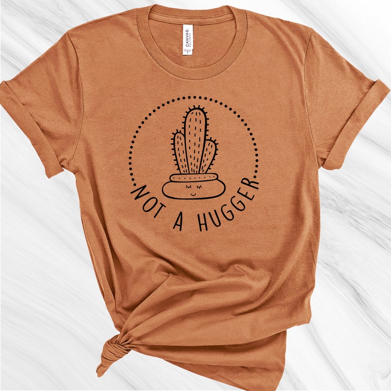 Puede incluir: Una camiseta marr&oacute;n con un gr&aacute;fico de cactus negro y el texto "Not a Hugger" en un c&iacute;rculo.