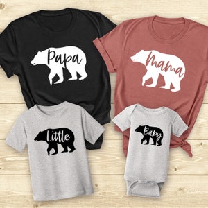 May include: A set of four t-shirts featuring a bear silhouette with the words "Papa", "Mama", "Little", and "Baby" printed on them. The shirts are in black, maroon, and gray.