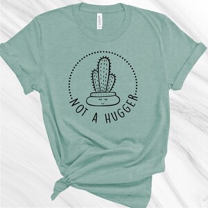 Puede incluir: Una camiseta verde claro con un gr&aacute;fico de cactus negro y el texto "Not a Hugger" en un c&iacute;rculo.