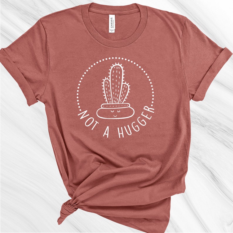Puede incluir: Una camiseta color marr&oacute;n rojizo con un gr&aacute;fico de cactus blanco y el texto "Not a Hugger" en un c&iacute;rculo.