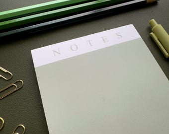 Puede incluir: Un bloc de notas verde con la palabra "NOTES" impresa en blanco en la parte superior. El bloc de notas está sobre una superficie verde con dos lápices verdes y un clip de papel dorado.
