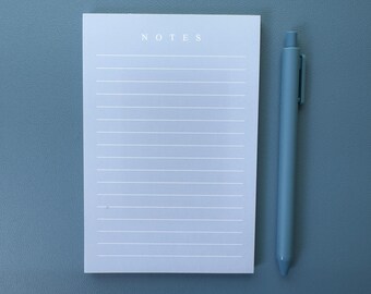 Puede incluir: Un bloc de notas blanco con la palabra "NOTES" impresa en la parte superior. El bloc de notas tiene papel rayado y está sobre una superficie azul. Un bolígrafo azul está al lado del bloc de notas.