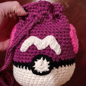 May include: Purple and pink crocheted Pokeball with a white 'M' on the front. The Pokeball is a popular character from the Pokemon franchise.