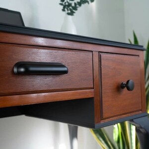 May include: A wooden desk with black painted legs and trim. The desk has two drawers with black hardware. The top drawer has a long black pull handle, and the bottom drawer has a round black knob.