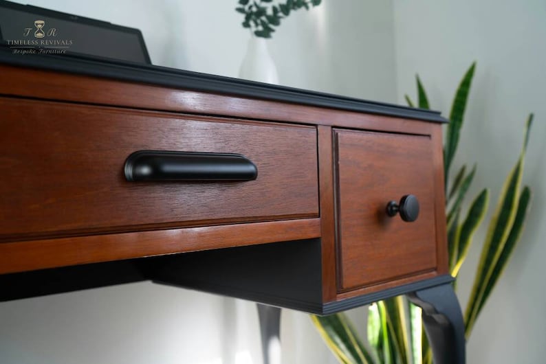 May include: A wooden desk with black painted legs and trim. The desk has two drawers with black hardware. The top drawer has a long black pull handle, and the bottom drawer has a round black knob.