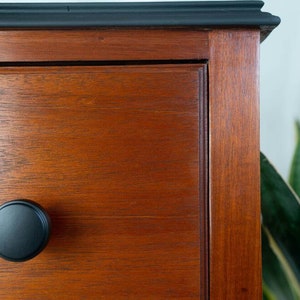 May include: Close-up of a wooden dresser drawer with a black knob. The drawer is made of a rich, dark wood with a smooth finish. The knob is round and has a simple design.