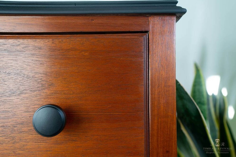 May include: Close-up of a wooden dresser drawer with a black knob. The drawer is made of a rich, dark wood with a smooth finish. The knob is round and has a simple design.