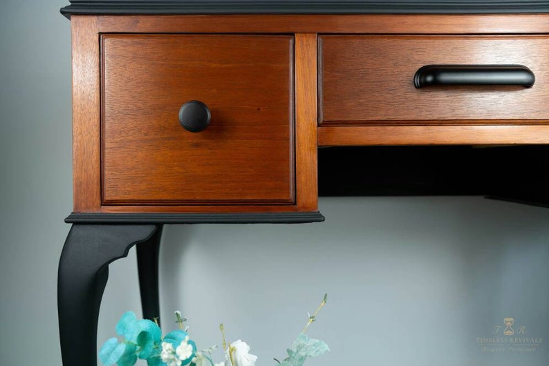 May include: A wooden desk with two drawers and black painted legs. The top of the desk is painted black, and the drawers are a natural wood color. The desk has a simple, elegant design.