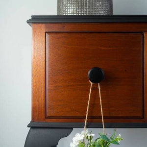 May include: A wooden dresser with black painted trim and a black knob. The dresser has two drawers and a small shelf. A small bouquet of white flowers hangs from the knob.