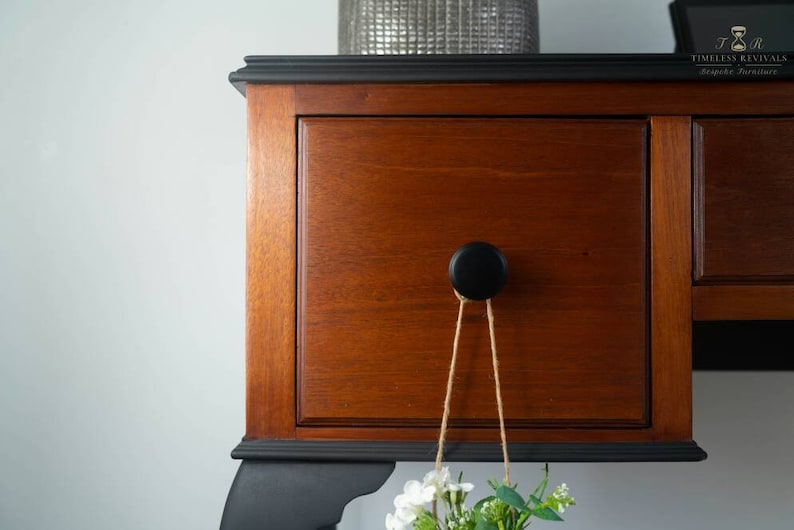 May include: A wooden dresser with black painted trim and a black knob. The dresser has two drawers and a small shelf. A small bouquet of white flowers hangs from the knob.