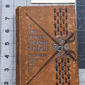 Peut inclure: Un livre relié vintage intitulé The Bow of Orange Ribbon par Amelia E. Barr avec une couverture brune, des illustrations décoratives d'épée et de ruban en noir et argent, et une note indiquant une largeur de dos de 1,9 cm sur un tapis de découpe.
