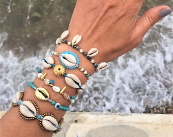 May include: A set of six bracelets featuring white seashells, turquoise beads, and gold accents. The bracelets are stacked on a person's wrist.