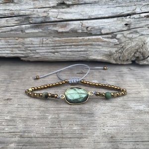 May include: A green labradorite gemstone bracelet with gold beads and a gray cord. The bracelet is on a wooden surface.