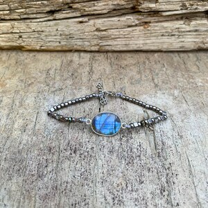 Puede incluir: Un brazalete de plata con una piedra preciosa de labradorita azul y cuentas de hematites negras.
