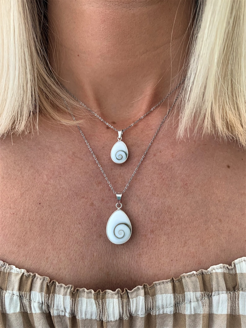 May include: Two silver necklaces with a white shell pendant featuring a green spiral design. The necklaces are layered on a person's neck.