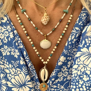 Peut inclure: Un collier à plusieurs rangs avec des perles blanches et turquoise et trois pendentifs en forme de coquillages. Le collier présente un grand pendentif en forme de coquillage blanc, un plus petit pendentif en forme de coquillage blanc et un pendentif en forme de coquillage brun.
