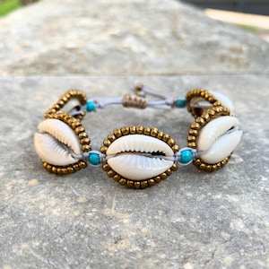 Può includere: Un braccialetto fatto a mano con conchiglie bianche bordate di perline color oro. Le conchiglie sono collegate da un cordoncino grigio, con piccole perline turchesi. Un accessorio elegante.