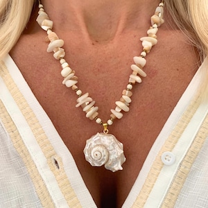 Op de afbeelding: Een ketting met een schelpenhanger. De ketting heeft een grote, witte schelpenhanger die hangt aan een streng kleine, onregelmatige, gebroken witte kralen en goudkleurige afstandhouders. De ketting wordt gedragen over een wit overhemd.