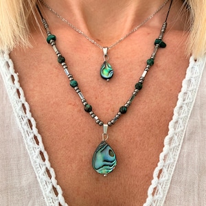 May include: Two necklaces with abalone shell pendants. The top necklace is a silver chain with a smaller abalone shell pendant. The bottom necklace is a beaded chain with a larger abalone shell pendant. The beads are green and silver.