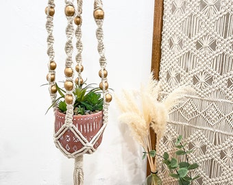 May include: A macrame plant hanger with gold beads and a terracotta pot with a succulent plant. The hanger is hanging from a ceiling hook and is in front of a white wall. There is a small table with a book, a candle, and a vase of dried flowers.
