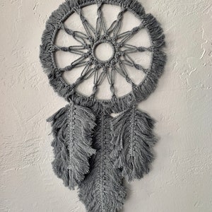 May include: A gray macrame dream catcher with a circular design and three long, fringed feathers hanging below.