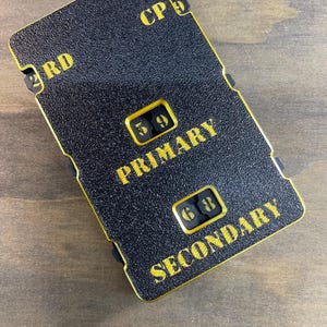 May include: A rectangular black textured device with gold edges and lettering. It features the text 2RD, CP 9, PRIMARY with the numbers 5 9, and SECONDARY with the numbers 6 8. The device has a structured, industrial appearance with clear labels.