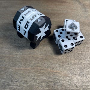 May include: A black and white dice roller with numbers and a star design, next to a pile of white dice with black pips and a spade symbol. The dice are on a wooden surface.