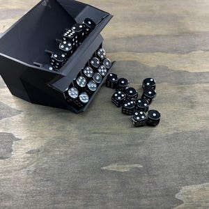 May include: A black dice tray with a geometric design, filled with black dice with white dots. Some dice are spilling out onto a wooden surface. The dice are standard six-sided dice, suitable for tabletop games.
