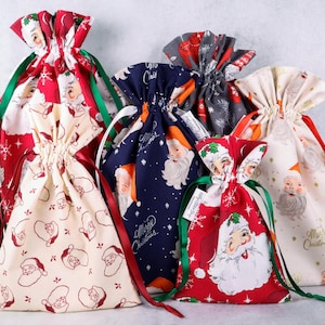 May include: A collection of five fabric drawstring gift bags featuring various Santa Claus prints in red, navy blue, and cream colors. Each bag is cinched at the top with satin ribbons in red or green, showcasing festive holiday patterns.