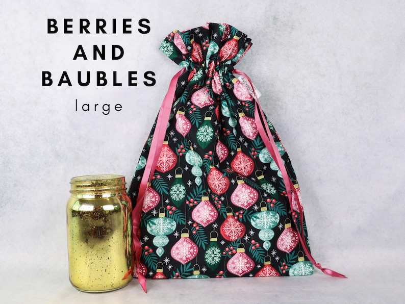 May include: A black fabric drawstring gift bag featuring a repeating pattern of red and teal Christmas ornaments, green pine needles, and red berries. The bag has pink ribbon ties and the text BERRIES AND BAUBLES large. A gold mercury glass jar.