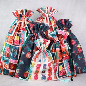 May include: A collection of five fabric drawstring gift bags in varying sizes. The bags feature colorful holiday-themed patterns including houses, candy, and ornaments in shades of red, green, blue, and cream with green ribbon ties.