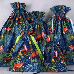 May include: A collection of drawstring bags with a repeating forest and fox pattern. The bags are navy blue with orange foxes, green foliage, and yellow flowers. The bags have ribbons in various colors.