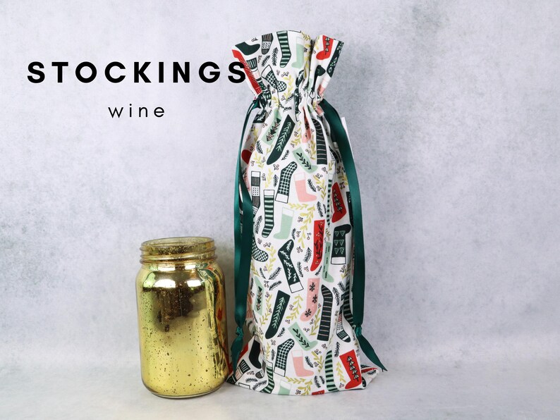 May include: A fabric wine bottle gift bag featuring a repeating pattern of colorful Christmas stockings in red, green, and black on a white background, cinched with a dark green ribbon. Next to it sits a gold-colored glass jar. Text reads STOCKINGS wine.