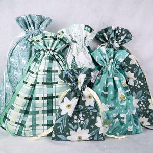 May include: A collection of six fabric drawstring gift bags in various sizes, featuring festive patterns in shades of green, white, and teal, including prints of deer, plaid, winter foliage, and cats wearing scarves with white ribbon ties.