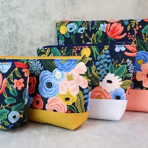 May include: A set of four zippered pouches in varying sizes, each featuring a dark navy blue fabric with a vibrant floral pattern of pink, blue, yellow, and white flowers. The pouches have solid-colored bases in mustard yellow, white, and peach.