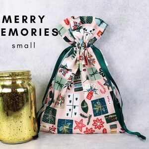 May include: A small fabric drawstring gift bag with a light pink background featuring a pattern of holiday presents, candy canes, and stars, paired with a gold mercury glass jar. The text MERRY MEMORIES small is visible.