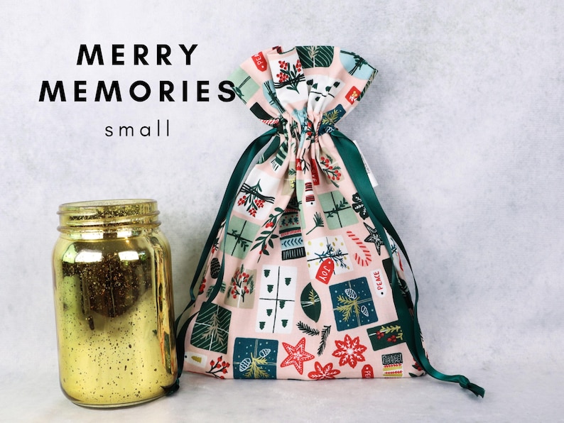May include: A small fabric drawstring gift bag with a light pink background featuring a pattern of holiday presents, candy canes, and stars, paired with a gold mercury glass jar. The text MERRY MEMORIES small is visible.