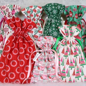 May include: A collection of seven fabric drawstring gift bags featuring various holiday-themed patterns including red wreaths, green snowflakes, pink gift boxes, and colorful Christmas trees, each finished with a matching satin ribbon tie.