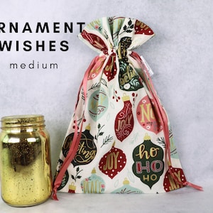 May include: A medium fabric gift bag with a drawstring closure, featuring a pattern of colorful Christmas ornaments with text including 'JOY', 'PEACE', and 'HO HO HO'. Beside it sits a gold-colored glass jar with a speckled finish.