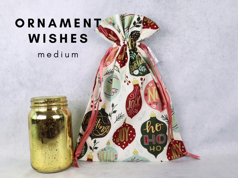 May include: A medium fabric gift bag with a drawstring closure, featuring a pattern of colorful Christmas ornaments with text including 'JOY', 'PEACE', and 'HO HO HO'. Beside it sits a gold-colored glass jar with a speckled finish.