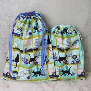 May include: Two blue and green drawstring bags with a black cat pattern. The larger bag has blue ribbon ties and the smaller bag has green ribbon ties.