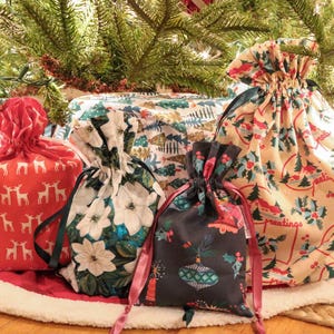 May include: A collection of four fabric drawstring gift bags featuring various holiday patterns including red with white reindeer, white with poinsettias, dark gray with ornaments, and cream with holly and the text greetings.