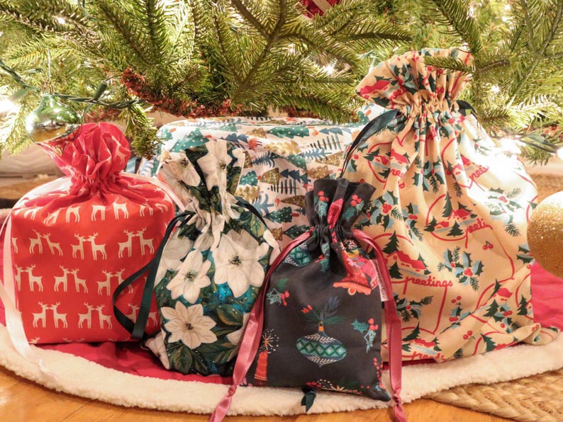 May include: A collection of four fabric drawstring gift bags featuring various holiday patterns including red with white reindeer, white with poinsettias, dark gray with ornaments, and cream with holly and the text greetings.
