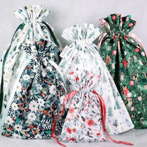 May include: Five fabric drawstring gift bags in varying sizes and patterns. Designs include white with green trees, dark green with cats and ornaments, dark blue with white flowers and red berries, and white with red poinsettias.