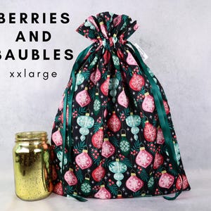 May include: A black fabric drawstring gift bag featuring a pattern of red, pink, and teal Christmas baubles, green pine needles, and red berries. The text BERRIES AND BAUBLES xxlarge is printed in black. A gold mercury glass jar sits to the left.