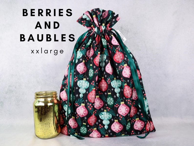 May include: A black fabric drawstring gift bag featuring a pattern of red, pink, and teal Christmas baubles, green pine needles, and red berries. The text BERRIES AND BAUBLES xxlarge is printed in black. A gold mercury glass jar sits to the left.