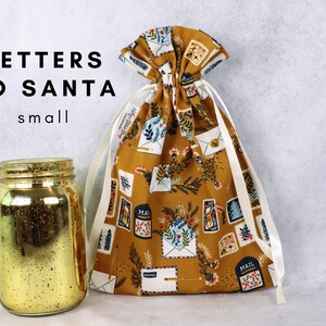 May include: A mustard yellow fabric drawstring bag featuring a pattern of envelopes, mailboxes, and pine branches, with white ribbon ties. Next to it sits a gold-colored glass jar. The text "LETTERS TO SANTA small" is visible in the upper left corner.