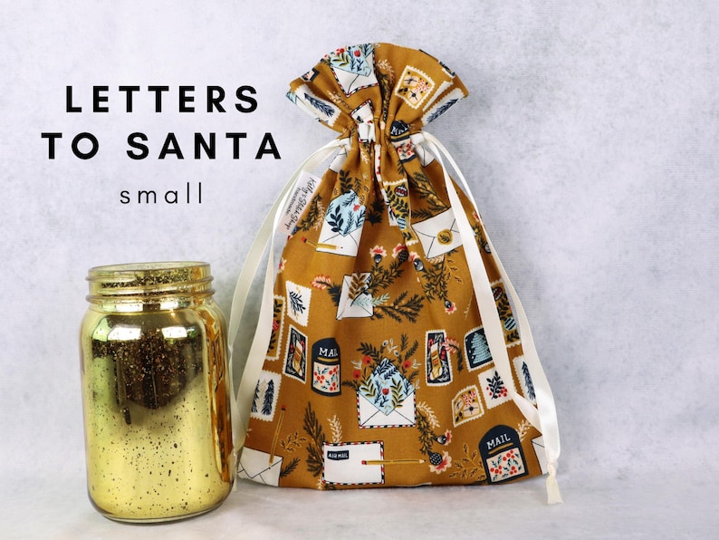 May include: A mustard yellow fabric drawstring bag featuring a pattern of envelopes, mailboxes, and pine branches, with white ribbon ties. Next to it sits a gold-colored glass jar. The text "LETTERS TO SANTA small" is visible in the upper left corner.