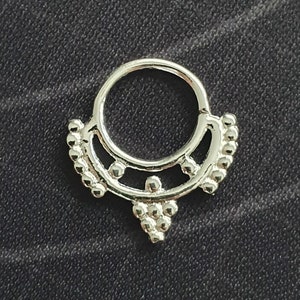 May include: Silver septum ring with a decorative design featuring a circle with small beads around the edge and a teardrop shape with beads hanging from the bottom.