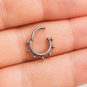 May include: A silver septum ring with small, evenly spaced ball accents. The hinged ring is open, showcasing the design. The jewelry is displayed on a light background.